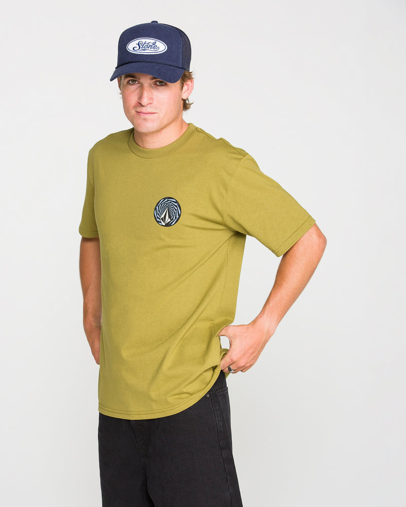 A person stands against a plain white background, hands on hips and looking at the camera, wearing a navy cap, black pants, and the Volcom Men's Dirty Work Short Sleeve Tee in olive green—a classic fit T-shirt featuring a circular logo on the chest. Crafted from premium ring-spun cotton, this tee offers both comfort and style.