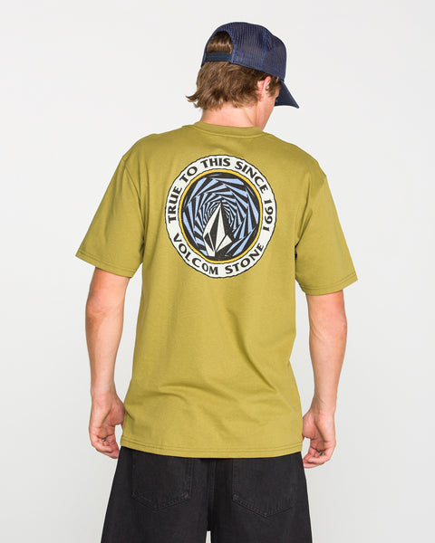 A man is seen from behind wearing the Volcom Men's Dirty Work Short Sleeve Tee in classic fit olive green, crafted from premium ring-spun cotton with a circular geometric logo and "True to This Since 1991 Volcom Stone" printed on the back. He pairs the tee with dark shorts and a navy cap.