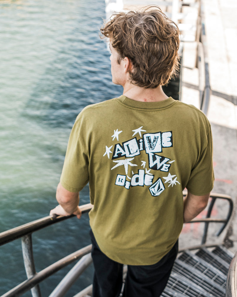 A person with curly hair stands on outdoor stairs by the water, wearing the Volcom Men’s Alive We Ride Short Sleeve Tee in olive green—crafted from heavyweight premium cotton and featuring bold ALiVE WE Ride graphic text. Sunlight casts shadows across the scene.