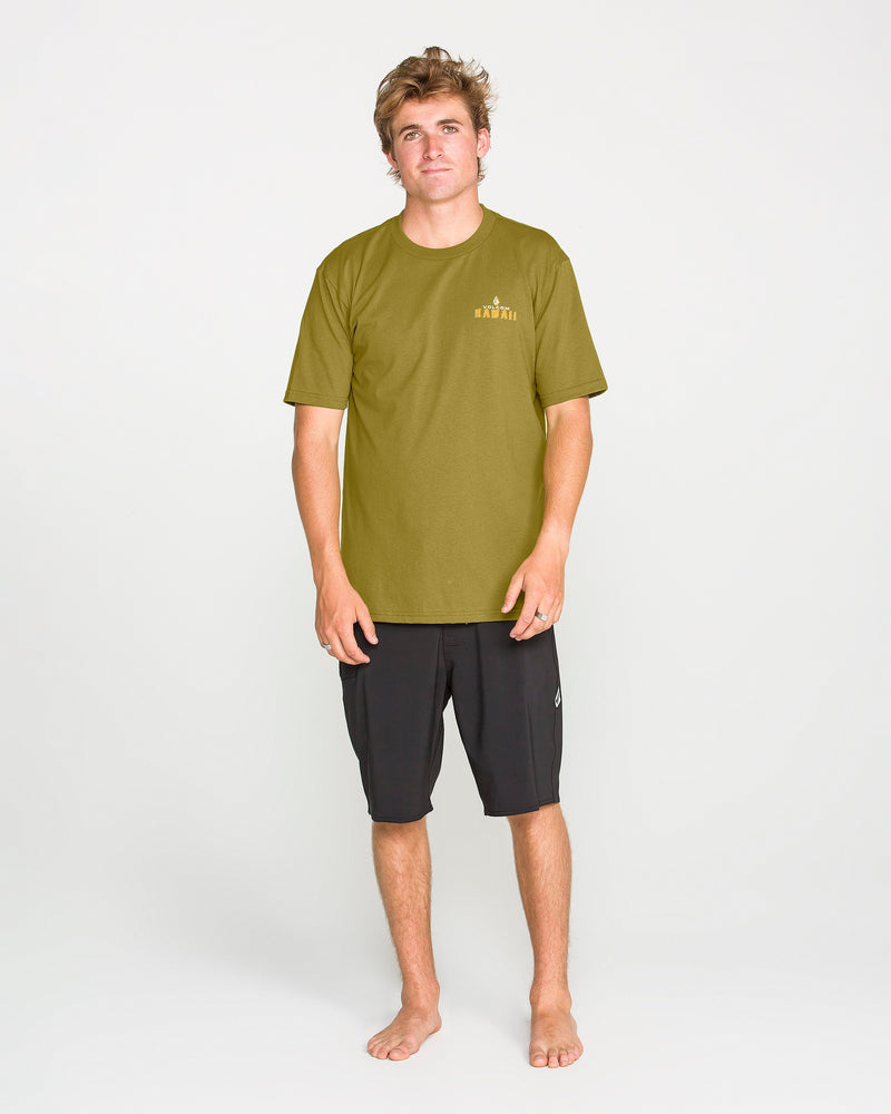 A man with short blond hair stands barefoot against a plain white background, wearing the Volcom Men’s Island Time Short Sleeve Tee in olive green, made from premium ring-spun cotton, along with black shorts. His arms rest relaxed by his sides as he faces forward with a neutral expression.