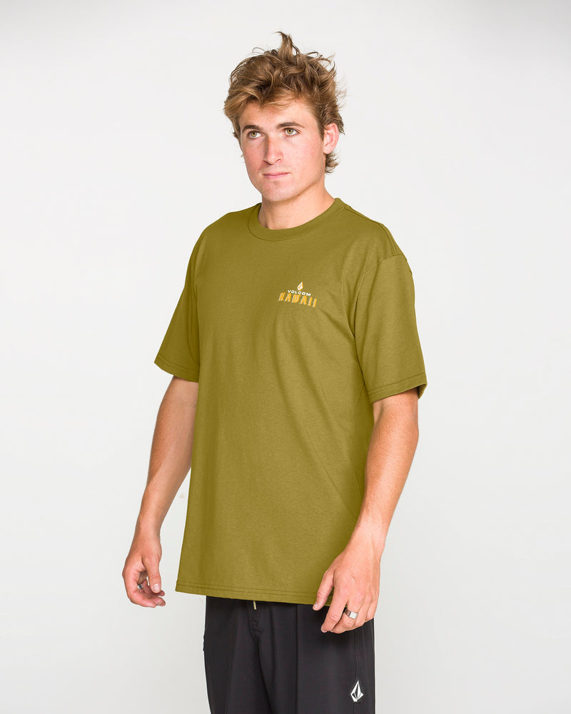 A young man with messy hair wears the Volcom Men’s Island Time Short Sleeve Tee in olive green, paired with black shorts, standing against a plain light gray background.