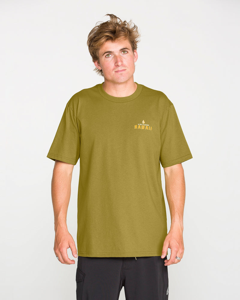 A young man with tousled hair wears the Volcom Men’s Island Time Short Sleeve Tee in olive green paired with black shorts, standing against a plain white background and looking at the camera with a neutral expression.