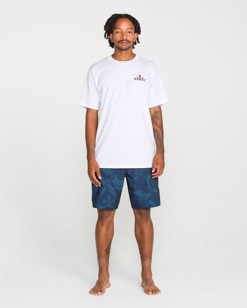 A man with short dreadlocks stands barefoot on a light background, wearing the Volcom Men’s Island Time Short Sleeve Tee—crafted from premium ring-spun cotton with a small chest graphic—and navy blue patterned shorts. His arms hang by his sides.