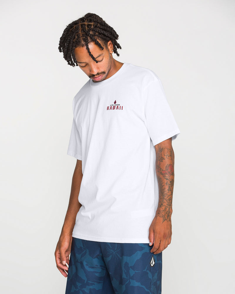 A man with short dreadlocks and tattoos on his left arm wears the Volcom Men’s Island Time Short Sleeve Tee made from premium ring-spun cotton, featuring HAWAII text, paired with blue camouflage shorts. He looks down with a neutral expression against a plain background.
