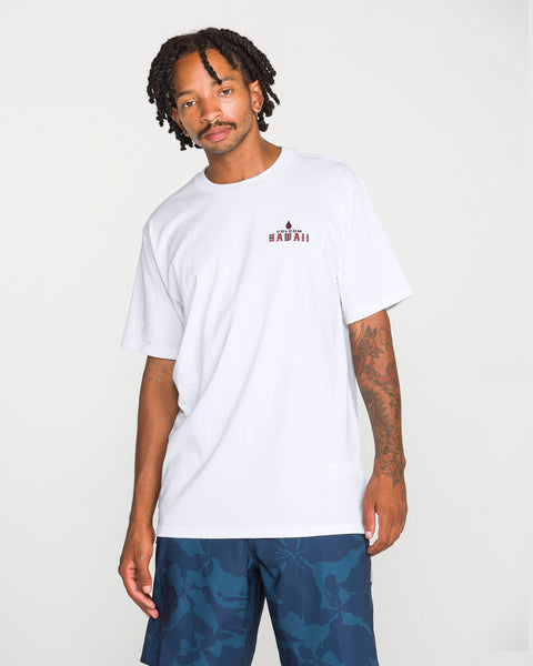 A man with short dreadlocks is wearing the Volcom Men’s Island Time Short Sleeve Tee, a classic fit t-shirt made from premium ring-spun cotton with small red graphic text on the chest, paired with blue patterned shorts and standing against a plain white background.