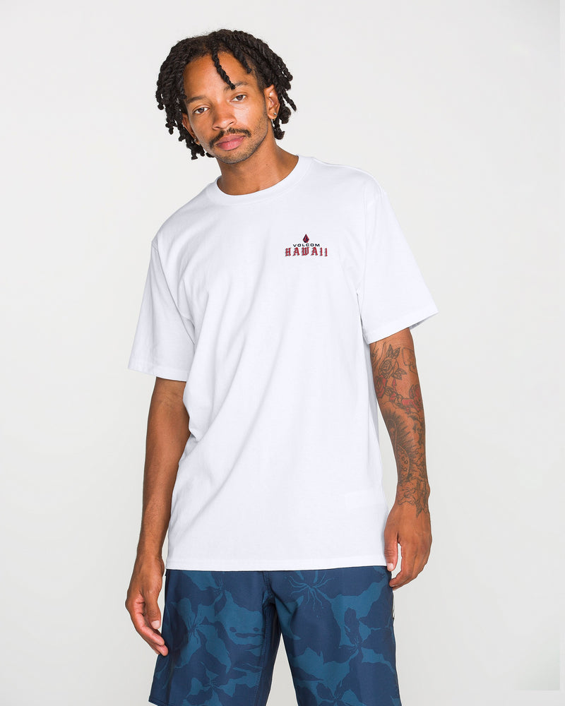 A man with short dreadlocks is wearing the Volcom Men’s Island Time Short Sleeve Tee, a classic fit t-shirt made from premium ring-spun cotton with small red graphic text on the chest, paired with blue patterned shorts and standing against a plain white background.