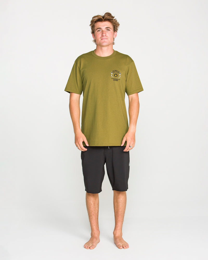 A person stands barefoot against a plain white background, wearing the Volcom Men’s Jungle Juice Short Sleeve Tee in green—a classic fit shirt with a small logo on the left chest, crafted from premium ring-spun cotton—and black knee-length shorts.