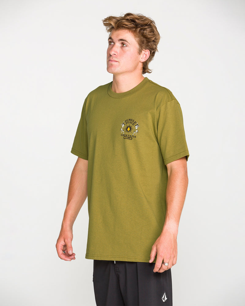 A young man stands against a neutral background, looking up while wearing the Volcom Men’s Jungle Juice Short Sleeve Tee in olive green—a classic fit made from premium ring-spun cotton with a small chest graphic—paired with black shorts.