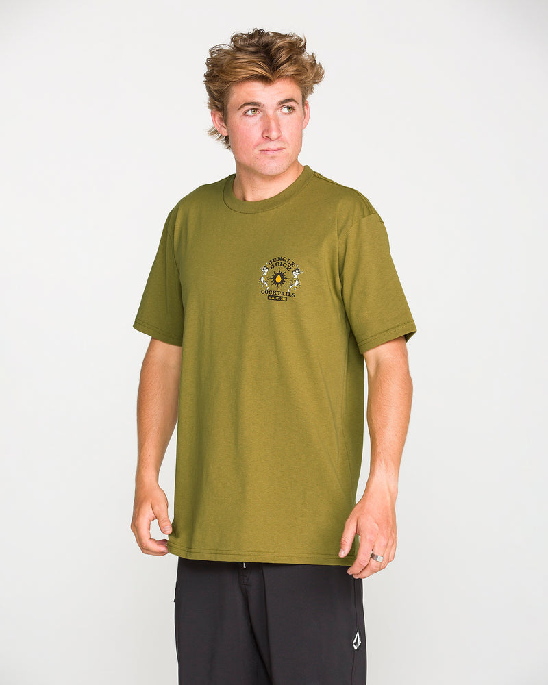 A person with light brown hair wearing the Volcom Men’s Jungle Juice Short Sleeve Tee in olive green premium ring-spun cotton and black pants stands against a plain white background, looking slightly to the side with a neutral expression.