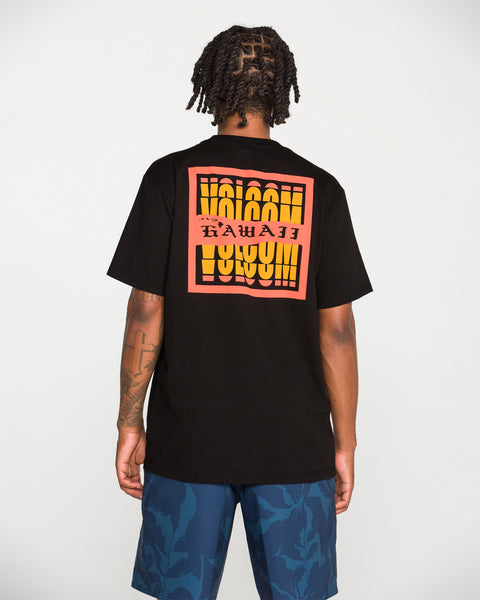 A person with short dreadlocks, seen from behind, wears Volcom’s Men's Refractor Short Sleeve Tee—a classic fit black T-shirt crafted from premium ring spun cotton and featuring a bold, colorful Hawaii graphic on the back—paired with blue patterned shorts, standing against a plain white background.