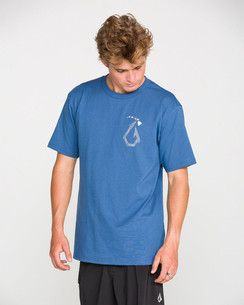 Wearing the Volcom Men’s Makau Short Sleeve Tee in blue, which features a small white graphic on the left chest, a person looks down while standing against a plain light background.