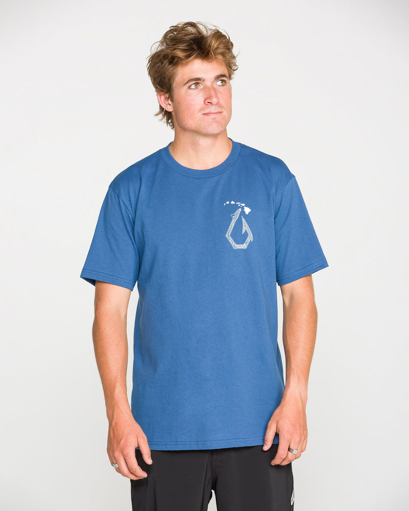 A young man wearing the Volcom Men’s Makau Short Sleeve Tee—crafted from heavyweight cotton in blue with a small abstract design on the left chest—stands against a plain white background, looking slightly upward and to the side.