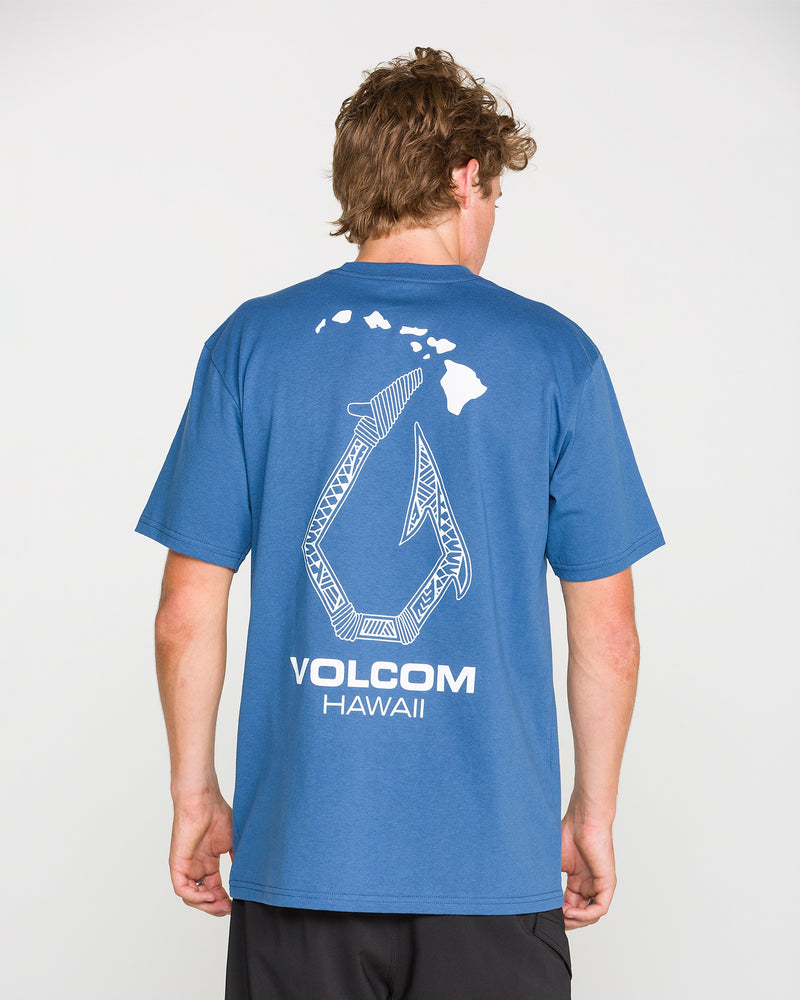 A person is seen from behind wearing the Volcom Men’s Makau Short Sleeve Tee, a blue heavyweight cotton t-shirt featuring a white graphic of a hook, the Hawaiian islands, and “VOLCOM HAWAII” text on the back.