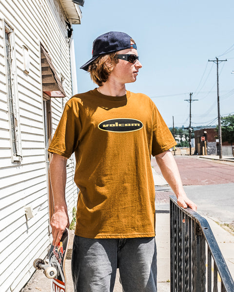 A young person in a Volcom Men's Quone Short Sleeve Tee, gray pants, and a backwards cap stands with a skateboard by a metal railing on a sunny sidewalk next to a white building.