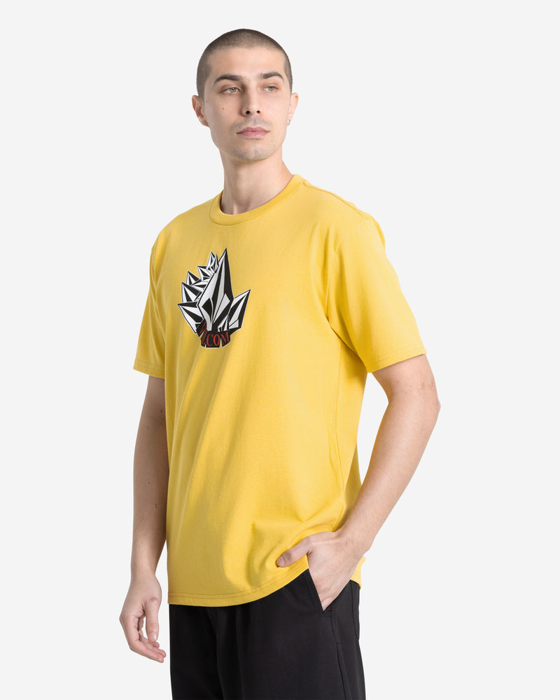 Wearing the Volcom Men's Shmoot Short Sleeve Tee in dark yellow with a black and white geometric graphic, and black pants, a person stands with one hand in their pocket, looking slightly to the side.