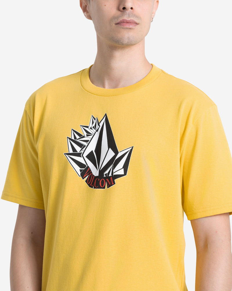 A person wears the Volcom Men's Shmoot Short Sleeve Tee, a dark yellow cotton t-shirt featuring an abstract black-and-white geometric graphic and red VOLCOM text on the chest; their face is partially out of frame.