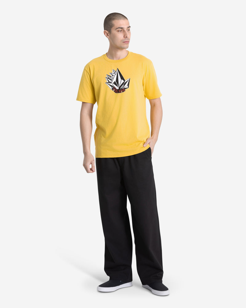 A man stands against a plain white background wearing the Volcom Men's Shmoot Short Sleeve Tee in dark yellow with a large black and white logo, paired with black wide-leg pants and black slip-on shoes.