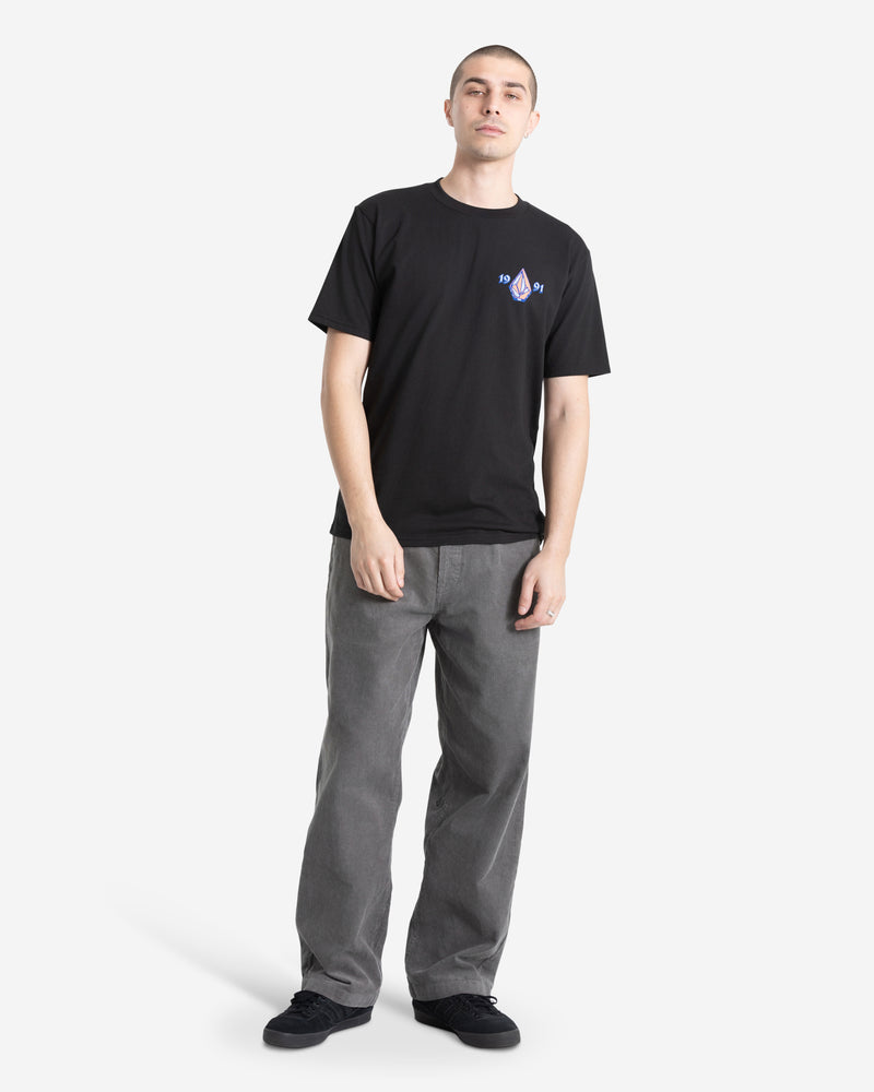 A person stands against a white background, wearing the Volcom Men's Countertop Short Sleeve Tee in black—100% cotton with a small chest graphic—paired with loose gray pants and black shoes. They have short hair and a relaxed expression, hands at their sides.