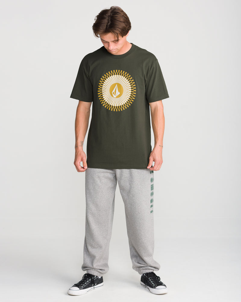 A person stands against a white background, wearing the Volcom Men’s Radiate Short Sleeve Tee, gray sweatpants, and black sneakers, looking down at the floor.