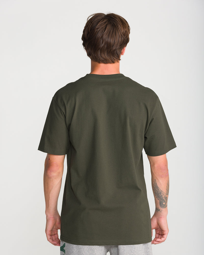 A person with short brown hair and a visible left forearm tattoo models the Volcom Men’s Radiate Short Sleeve Tee in olive green, paired with gray shorts, standing against a plain white background.