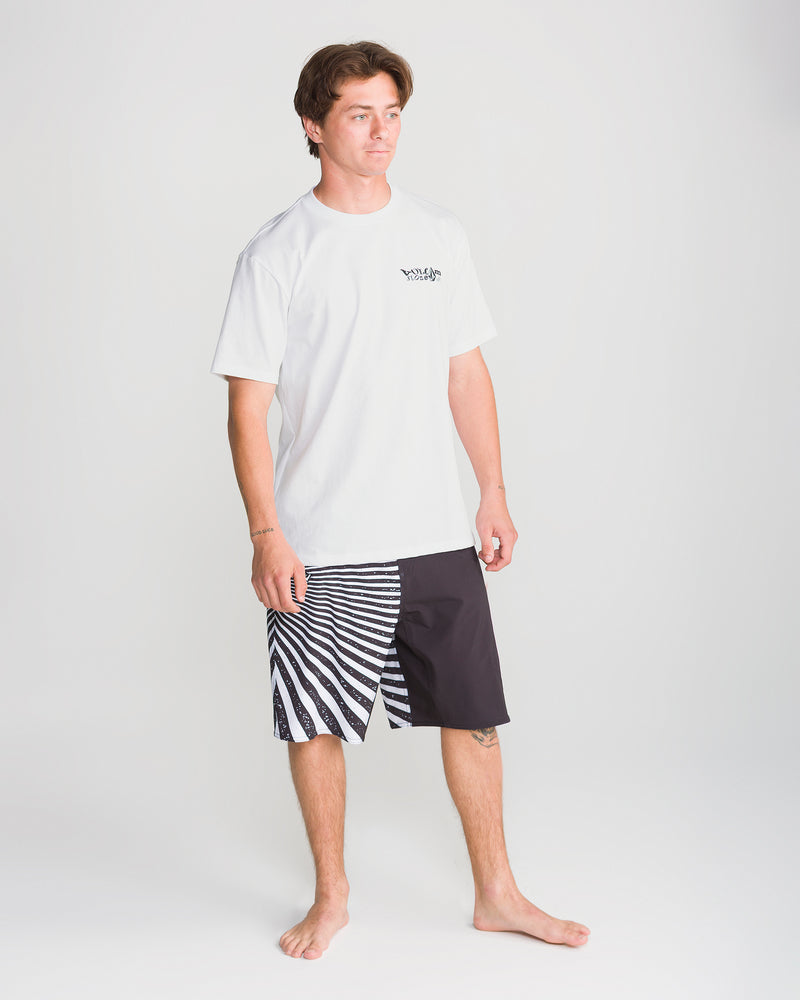 A young man stands barefoot on a white background, wearing the Volcom Men’s Framer Short Sleeve Tee and black shorts with a bold black-and-white zebra stripe pattern on one leg.