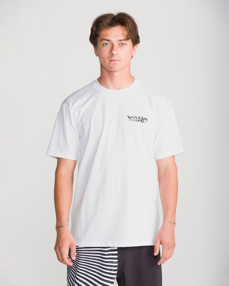 A young man with short brown hair wears the Volcom Men’s Framer Short Sleeve Tee, a classic fit white crew neck with a small black graphic on the left chest, paired with black-and-white striped shorts.