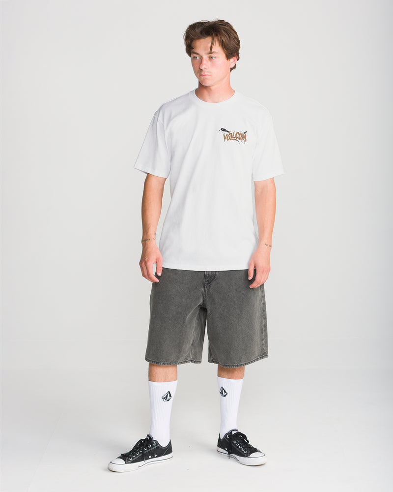 A young man stands against a plain background wearing the Volcom Men’s Zombeach Short Sleeve Tee, loose gray denim shorts, logo white socks, and black sneakers. He looks slightly to the side with a neutral expression.