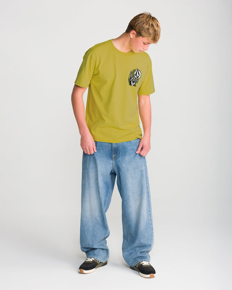 A person wearing the Volcom Men’s Opposition Short Sleeve Tee in yellow with a black and white graphic and wide blue jeans looks down against a plain white background.