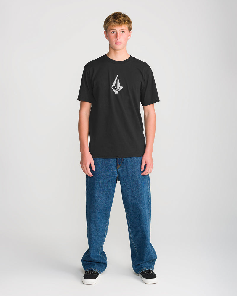 A young person stands facing forward, wearing the Volcom Men’s Full Stone Short Sleeve Tee in black—crafted from premium ring spun cotton and featuring a white geometric logo—paired with blue baggy jeans and black sneakers, set against a plain light background.