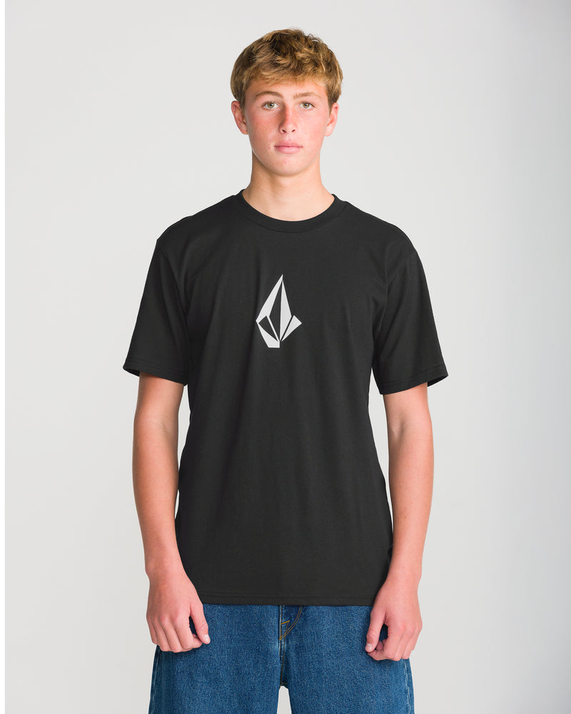 A young person stands facing forward, wearing the Volcom Men’s Full Stone Short Sleeve Tee—a black crew neck shirt featuring a geometric white logo on the chest—paired with blue jeans, against a plain light background.