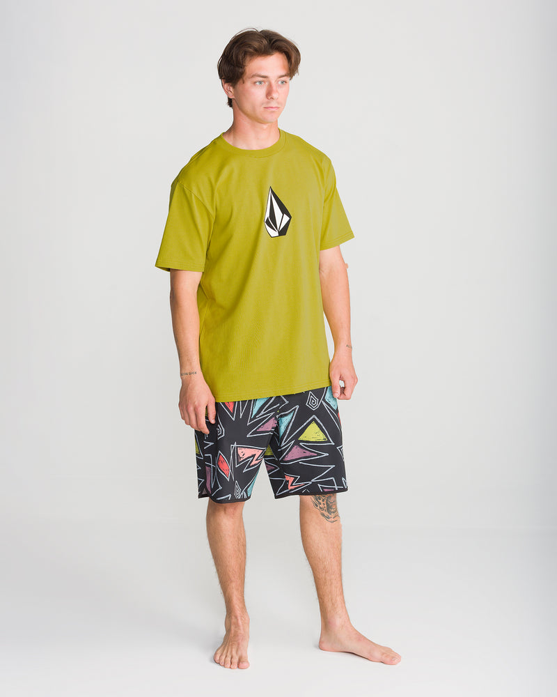 A young man stands barefoot in the Volcom Men’s Full Stone Short Sleeve Tee in yellow-green with a black and white logo, paired with black shorts featuring colorful abstract patterns, against a plain white background.