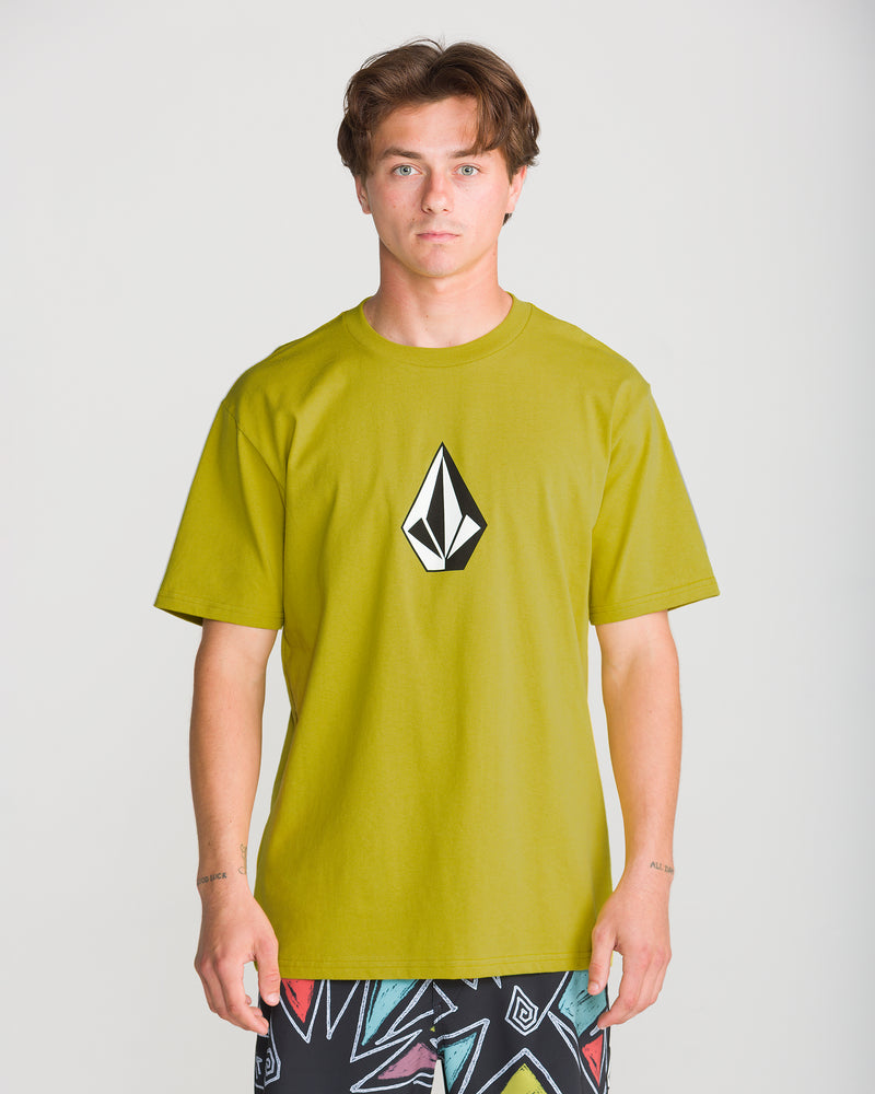A young man stands facing forward in a lime green Volcom Men’s Full Stone Short Sleeve Tee with a black and white logo, paired with patterned shorts against a plain light background.