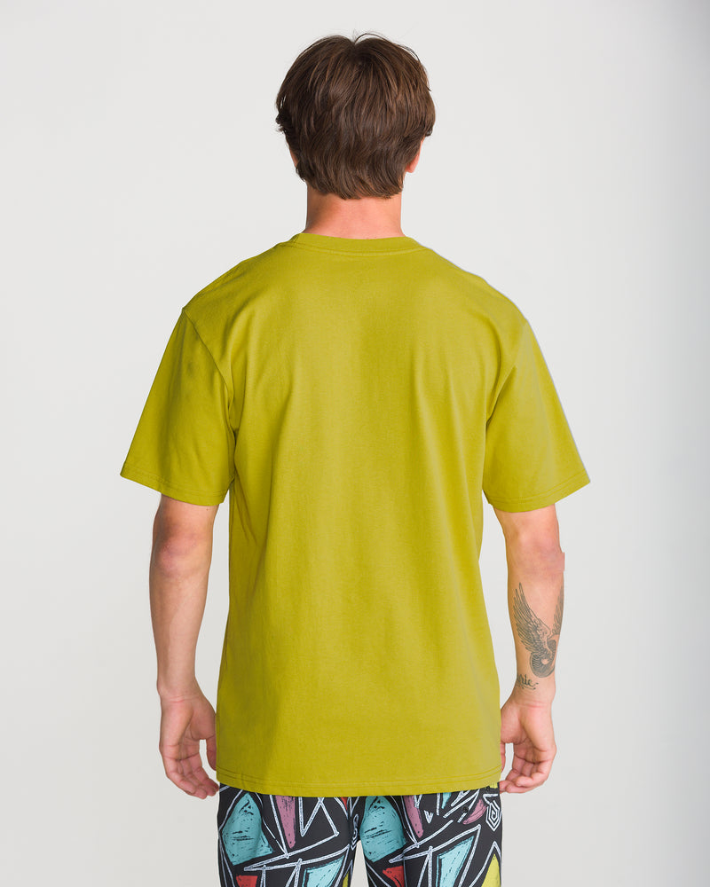 A person with short brown hair, tattooed left forearm, stands with their back to the camera in a Volcom Men’s Full Stone Short Sleeve Tee (bright green) and abstract patterned shorts against a plain white background.