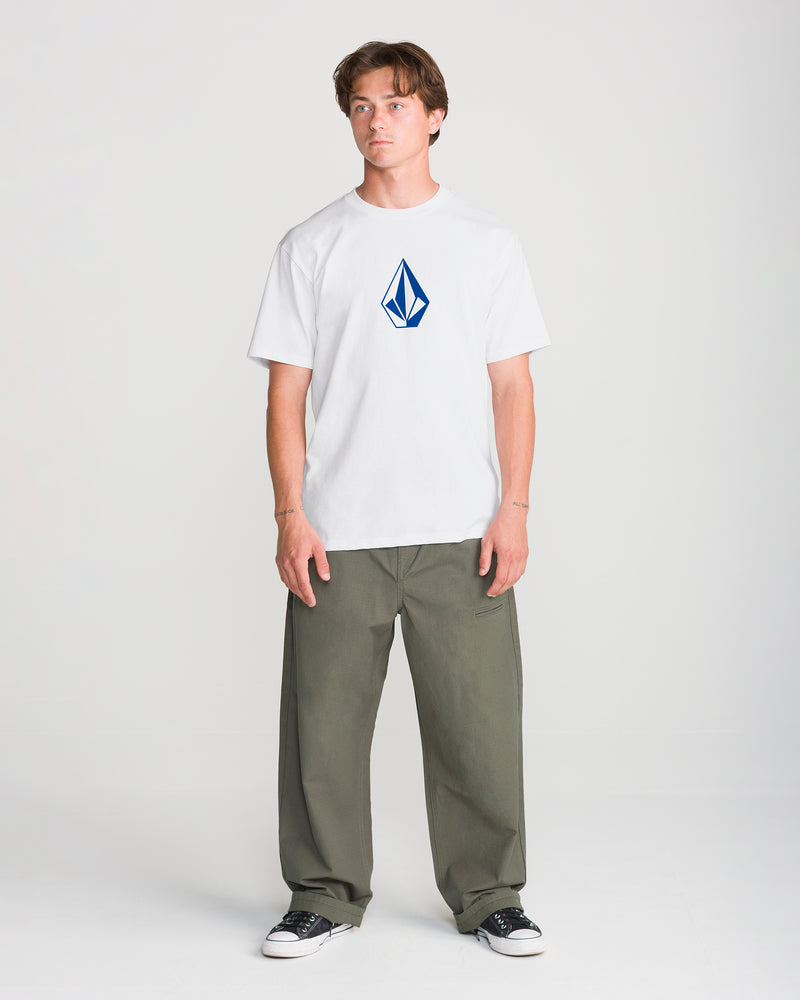 A person wearing a white Volcom Men’s Full Stone Short Sleeve Tee with a blue diamond-shaped logo on the chest, paired with olive green pants and black sneakers.