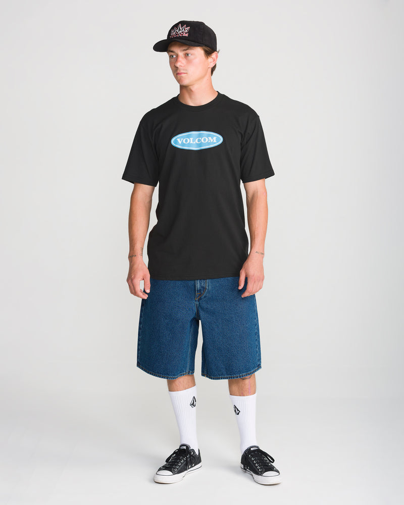 A man wears a black Men’s Remastered Stone Short Sleeve Tee with a blue oval logo on the chest, paired with denim shorts, white socks, and black sneakers.