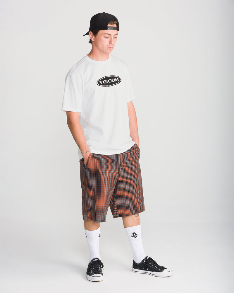 A man wearing a Volcom Men’s Remastered Stone Short Sleeve Tee with a black oval logo, plaid shorts, white crew socks, and black sneakers.