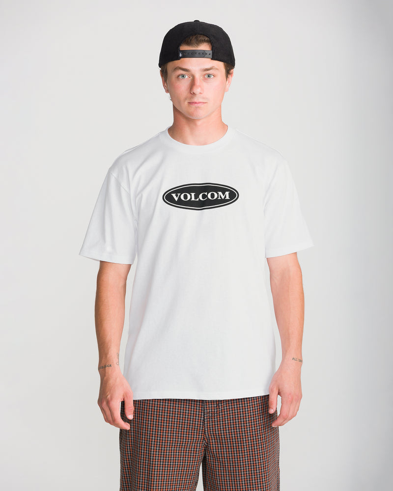 A person wearing a white Men’s Remastered Stone Short Sleeve Tee with a black oval logo, a black baseball cap, and plaid shorts.