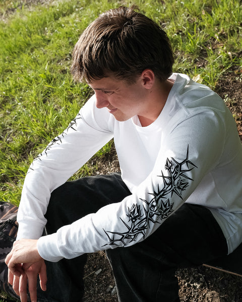 A young person with short brown hair sits outdoors on grass, wearing the Volcom Men's Die By The Stone Long Sleeve Tee featuring black graphic sleeve designs and dark pants, looking down with a faint smile.