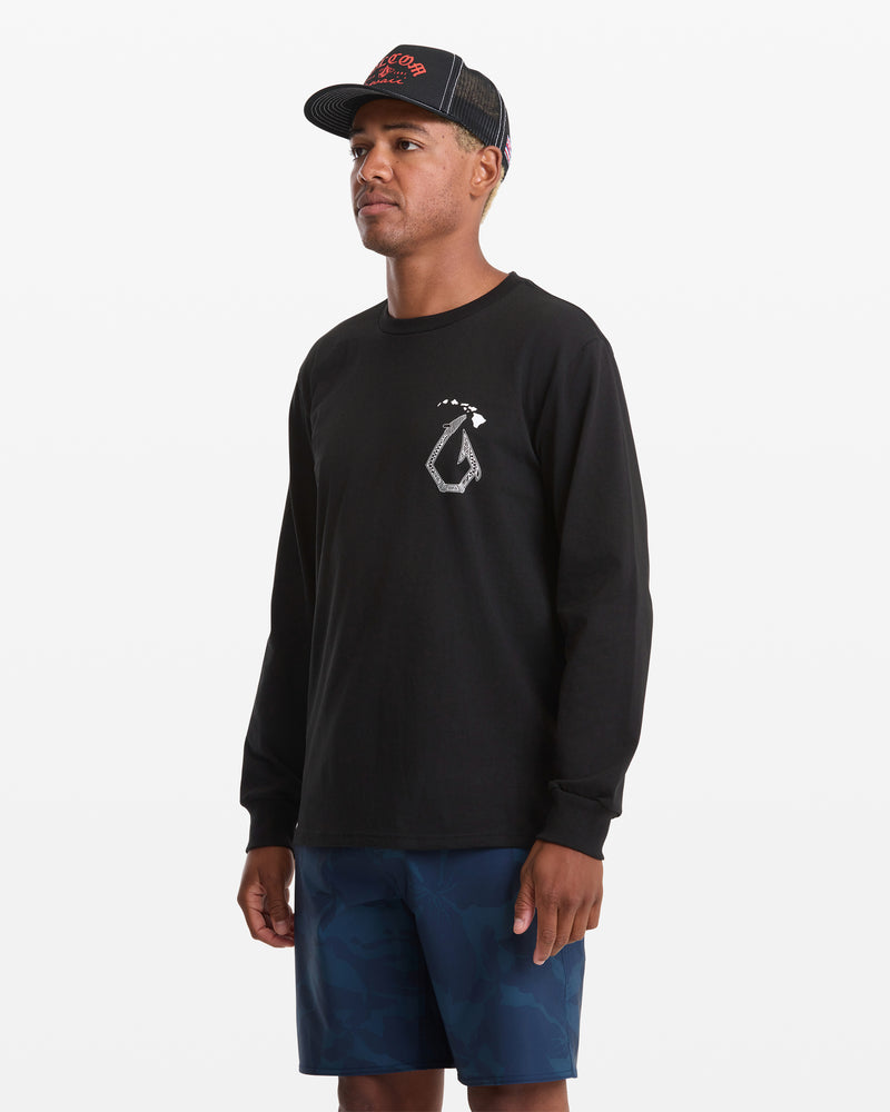 A man wears the Volcom Men's Makau Long Sleeve Tee in black with a white chest graphic, paired with blue camo shorts and a black mesh cap, standing against a plain white background.