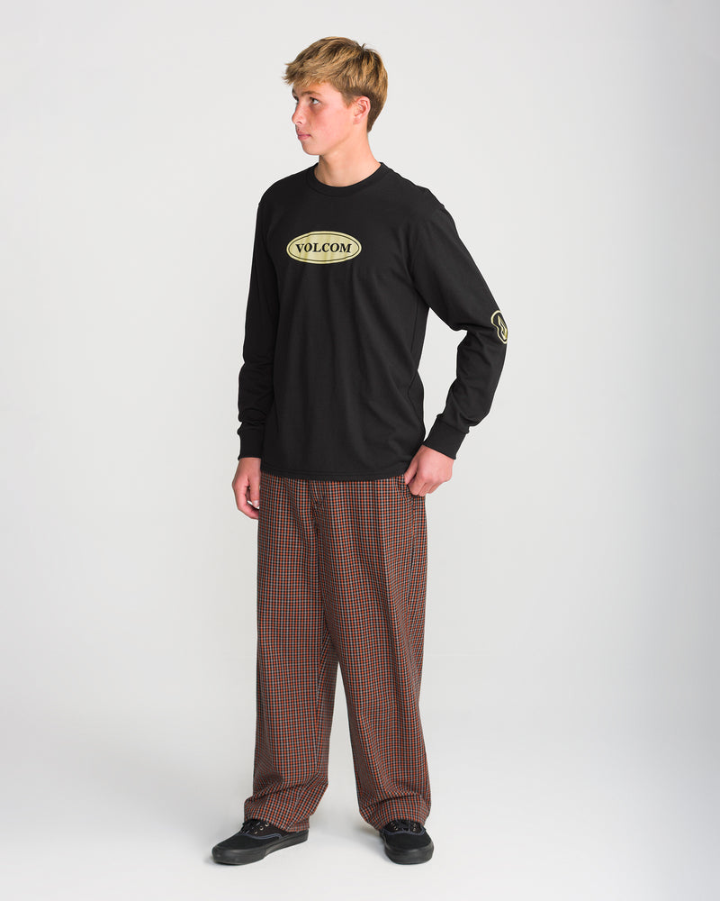 A person wears a black Volcom Men’s Remastered Stone Long Sleeve Tee featuring an oval chest logo and a sleeve graphic, paired with brown plaid trousers and black sneakers.