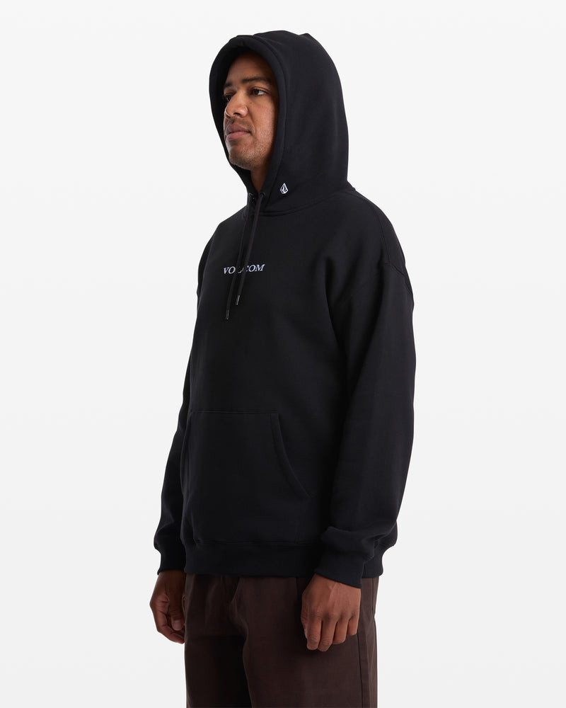 A man stands facing slightly left against a white background, wearing the Men's Volcom Stone Pullover—a black fleece hoodie by Volcom featuring VOLCOM on the front, made from recycled polyester and BCI cotton. Hood up, hands relaxed at his sides.
