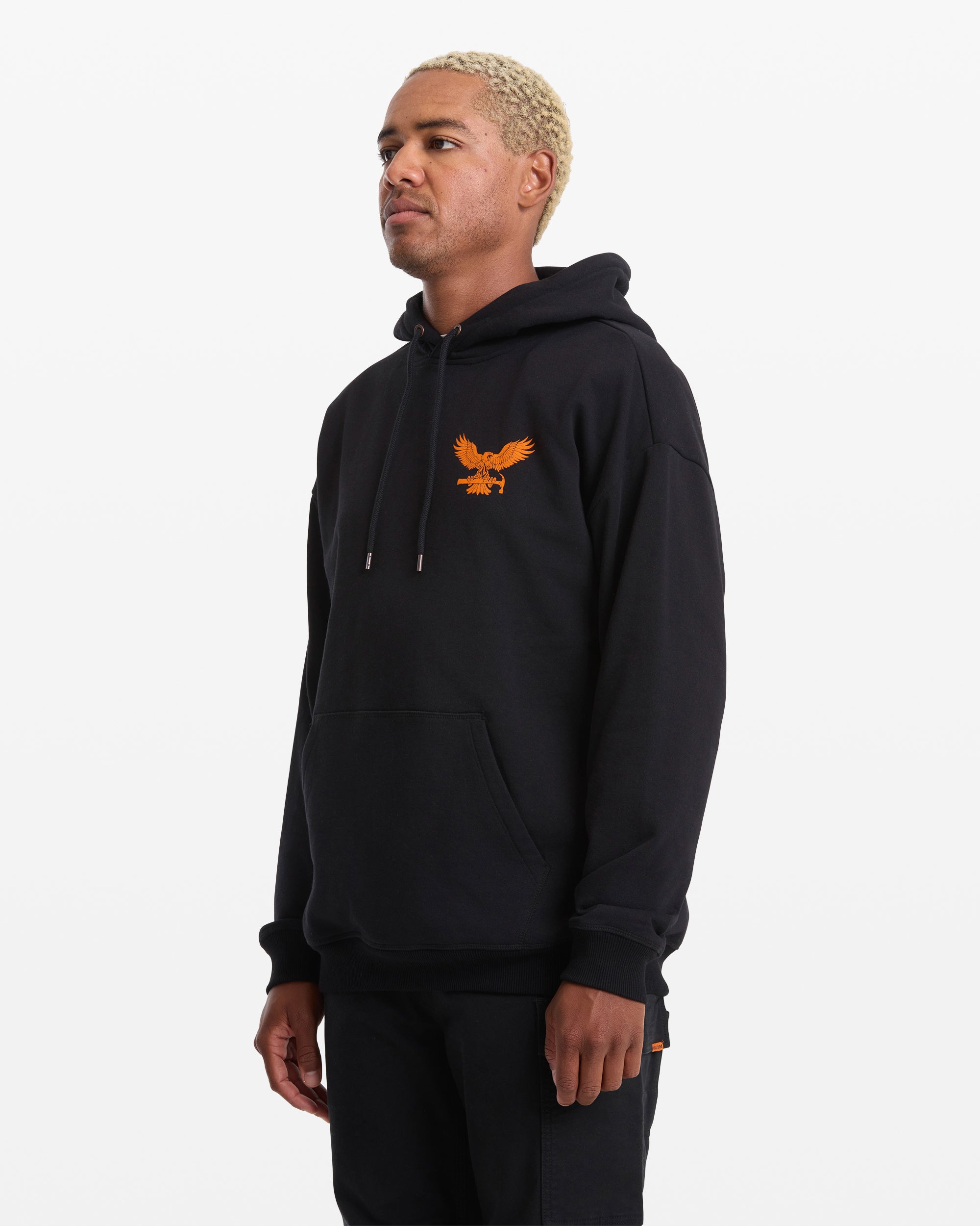 A person with short, light blonde hair wears the Volcom Men's Hammered Pullover—a black hoodie with an orange eagle graphic on the left chest. They stand against a plain white background, looking slightly to the side.
