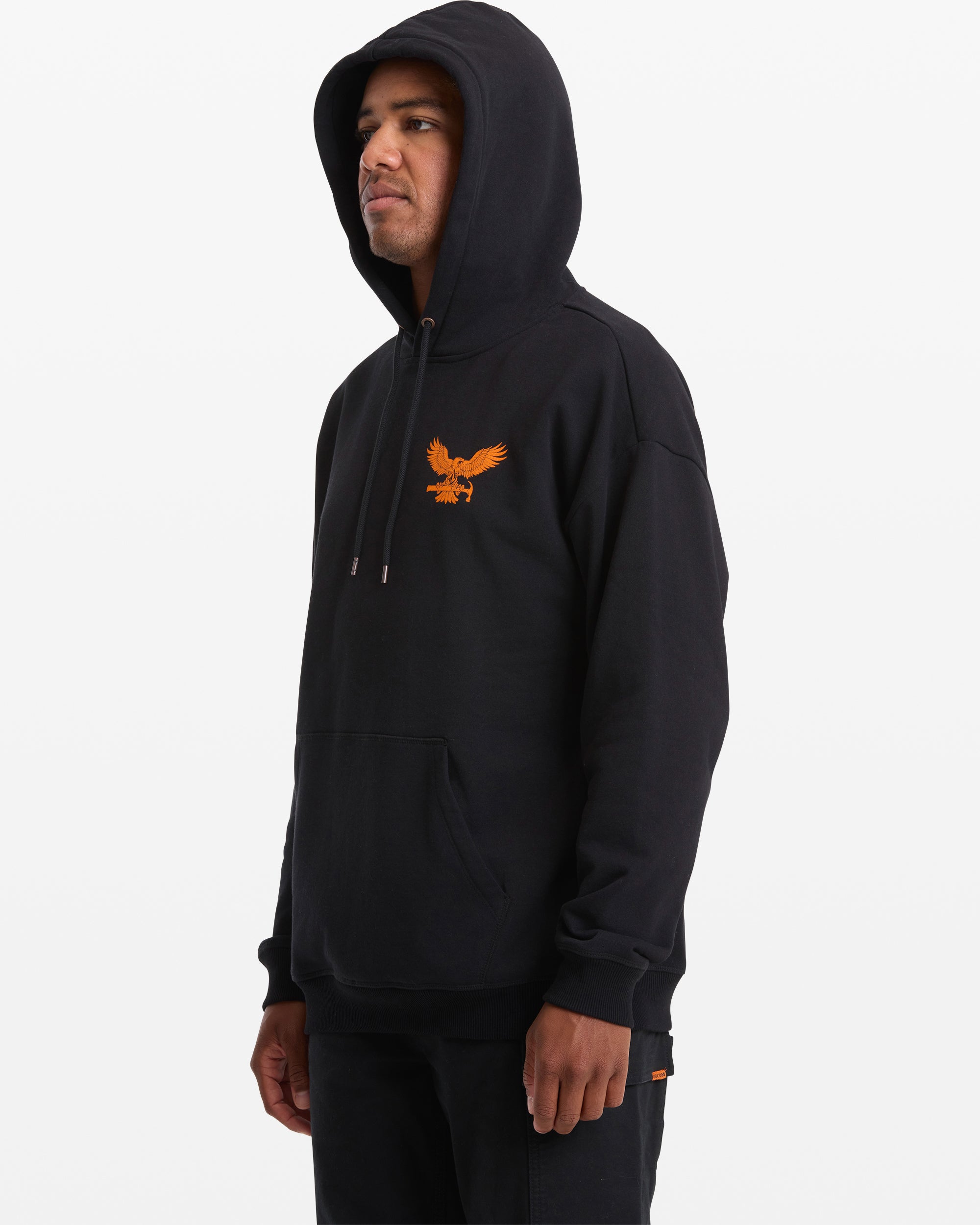 A man wears the Volcom Men's Hammered Pullover, a black hoodie with an orange eagle graphic on the left chest, and black pants, standing against a plain white background.
