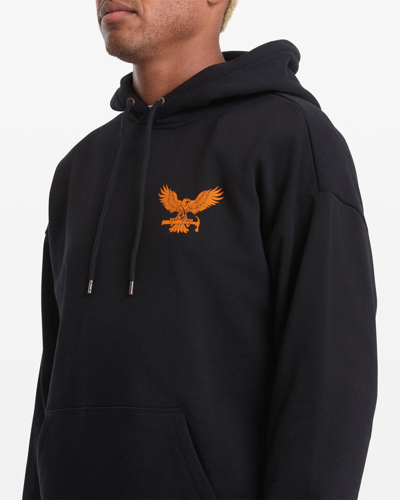 A person stands slightly to the side wearing the Volcom Men's Hammered Pullover, a black hoodie featuring an orange eagle graphic on the chest, front pocket, and drawstrings. The background is plain white.