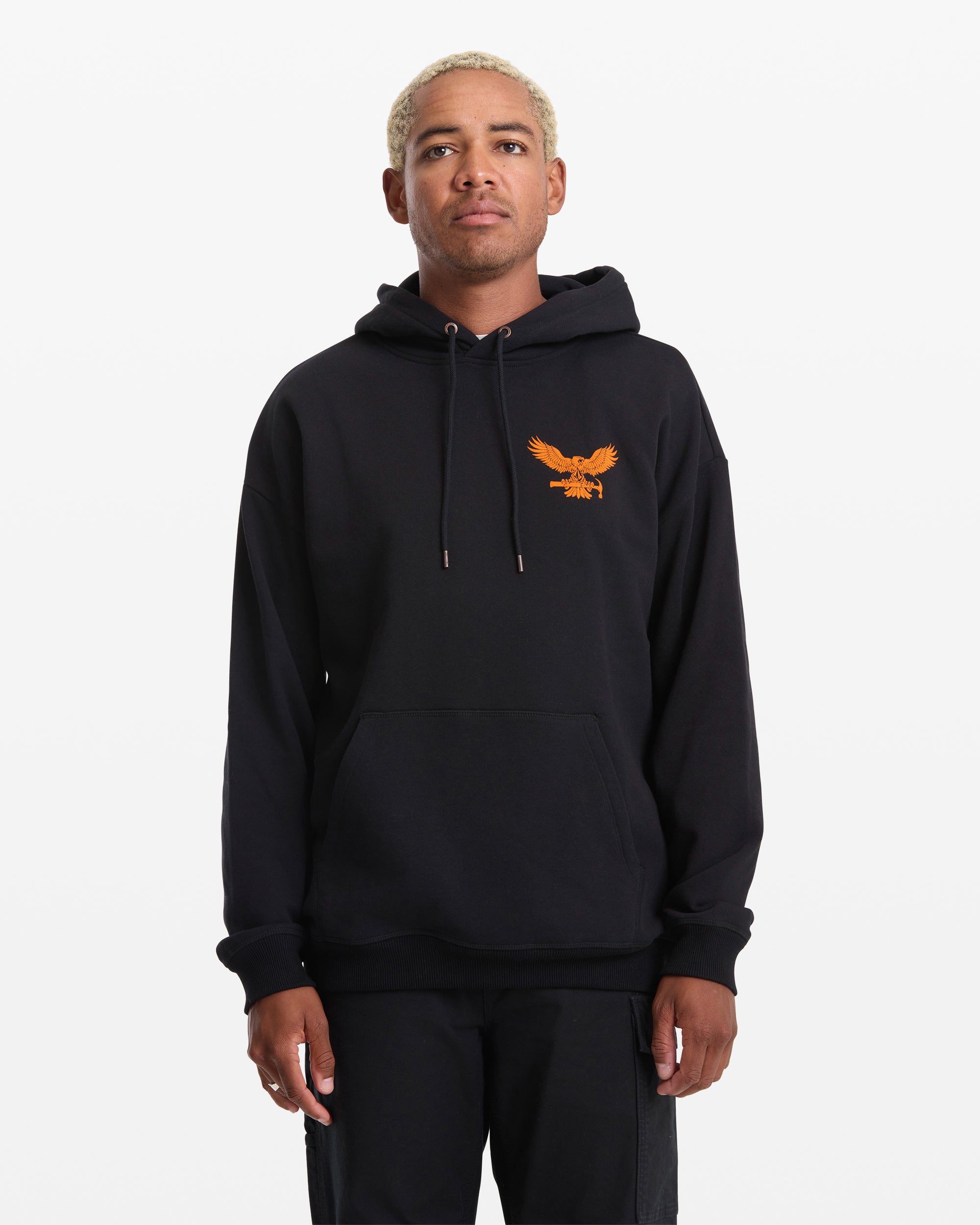A person with short blond hair wears the Volcom Men's Hammered Pullover, a black hoodie featuring an orange eagle graphic on the left chest, standing against a plain white background.