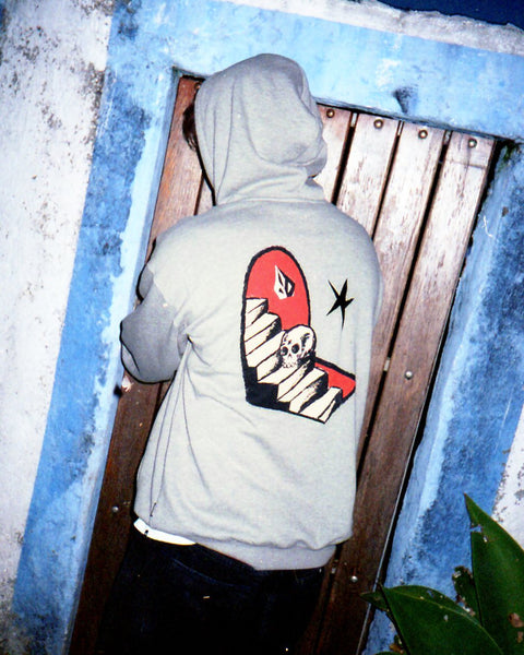 A person wearing the Volcom Men's Featured Artist Seth Conboy Pullover, which has a gravestone, steps, and skull graphic on the back, stands with their back to the camera and opens a wooden door framed by a blue-and-white wall.