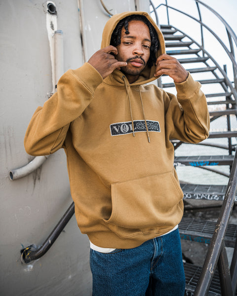 A person stands outdoors near a metal staircase, looking at the camera and adjusting their hood with one hand while wearing the Men’s Super Stoned Pullover by Volcom—crafted from cozy brushed-back fleece in tan—and pairing it stylishly with blue jeans.