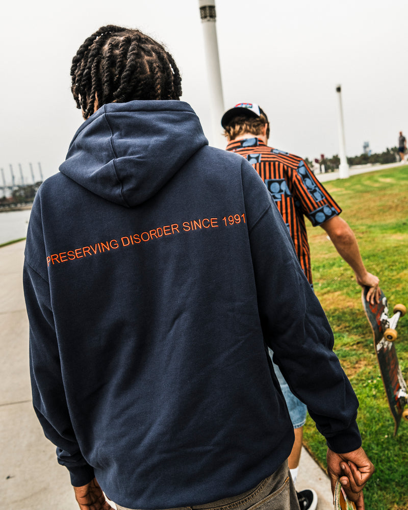 A person with braided hair wears the Volcom Men’s Featured Artist Antonio Carrau Pullover, a heavyweight fleece navy hoodie featuring “PRESERVING DISORDER SINCE 1991” in bold orange text, as they walk beside another person holding a skateboard in a park. Inspired by Antonio Carrau’s vibrant collages, this cozy pullover blends classic comfort with distinctive artistic flair.