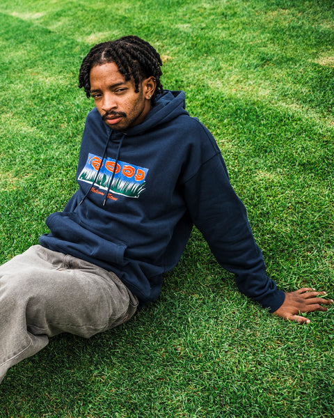 A man with short braids wearing the Volcom Men’s Featured Artist Antonio Carrau Pullover in navy and gray pants sits on green grass, looking at the camera with a neutral expression.