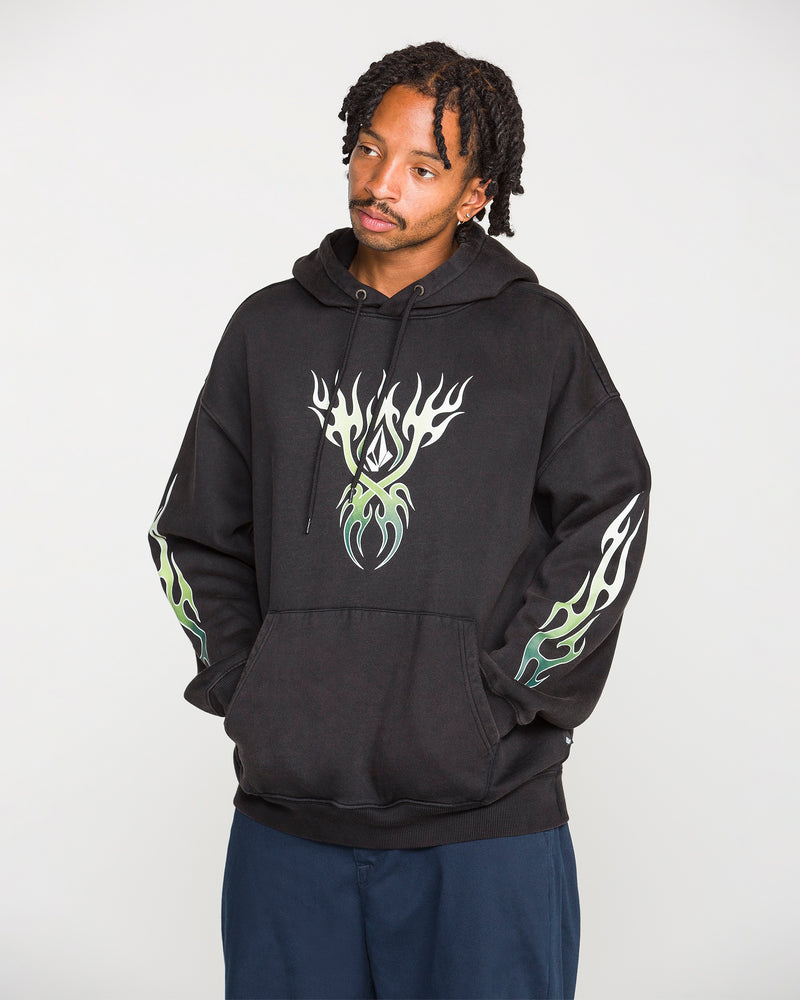 A person wearing the Volcom Men's Stone Trip Pullover—a washed-black hoodie featuring white and green flame designs on the chest and sleeves—stands with hands in the front pocket, looking slightly to the side.